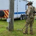 A fully equipped military or law enforcement operator in multicam camouflage is using a metal detector or ground-scanning device near a utility pole on a grassy area. The scene appears to be a forward operating base or staging area, with a white and red/yellow chevron-marked trailer, a tan shelter structure, and palletized water supplies visible in the background. The activity suggests an explosive ordnance disposal (EOD), route clearance, or security sweep operation.
