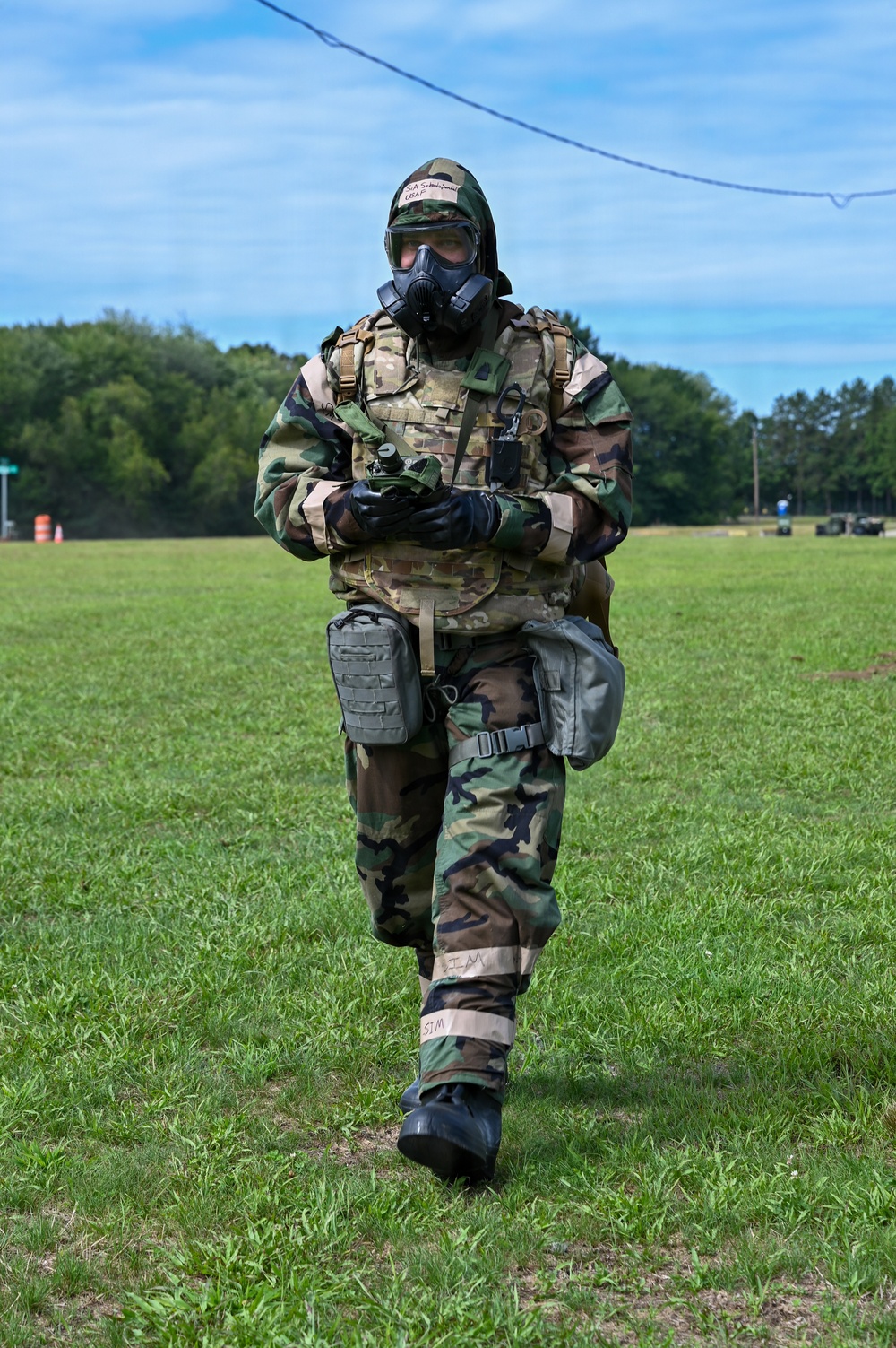 A military personnel wearing full CBRN (Chemical, Biological, Radiological, and Nuclear) protective gear walks across a grassy field during what appears to be a training exercise. The individual is equipped with a gas mask, woodland camouflage MOPP suit over a MultiCam tactical vest, black rubber gloves and boots, and carries various pouches and equipment. Tape markings labeled 'SIM' on the legs suggest this is a simulation or training scenario.