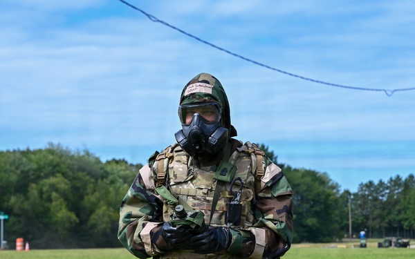 A military personnel wearing full CBRN (Chemical, Biological, Radiological, and Nuclear) protective gear walks across a grassy field during what appears to be a training exercise. The individual is equipped with a gas mask, woodland camouflage MOPP suit over a MultiCam tactical vest, black rubber gloves and boots, and carries various pouches and equipment. Tape markings labeled 'SIM' on the legs suggest this is a simulation or training scenario.