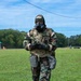 A military personnel wearing full CBRN (Chemical, Biological, Radiological, and Nuclear) protective gear walks across a grassy field during what appears to be a training exercise. The individual is equipped with a gas mask, woodland camouflage MOPP suit over a MultiCam tactical vest, black rubber gloves and boots, and carries various pouches and equipment. Tape markings labeled 'SIM' on the legs suggest this is a simulation or training scenario.