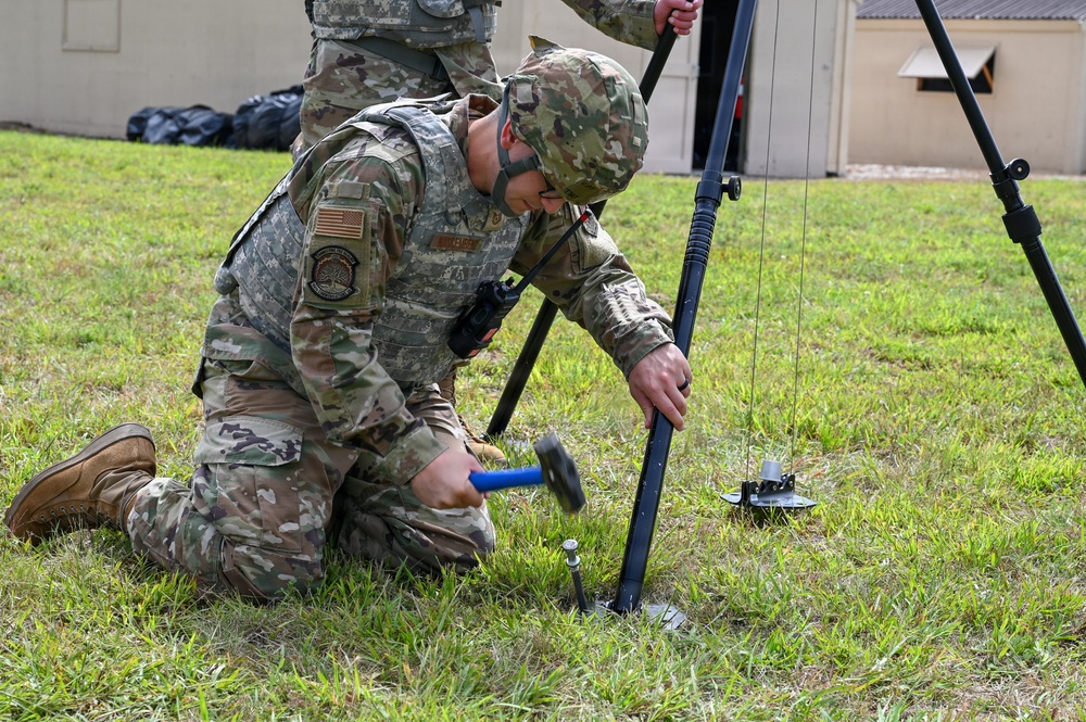 A U.S. Army soldier in OCP (Operational Camouflage Pattern) uniform is kneeling on grass, using a hammer to drive a ground stake into the earth as part of setting up a tripod-mounted antenna or communications mast. A second soldier stands behind, assisting with the upper portion of the equipment. The setting appears to be a training or operational area near a building.