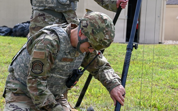 A U.S. Army soldier in OCP (Operational Camouflage Pattern) uniform is kneeling on grass, using a hammer to drive a ground stake into the earth as part of setting up a tripod-mounted antenna or communications mast. A second soldier stands behind, assisting with the upper portion of the equipment. The setting appears to be a training or operational area near a building.