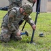 A U.S. Army soldier in OCP (Operational Camouflage Pattern) uniform is kneeling on grass, using a hammer to drive a ground stake into the earth as part of setting up a tripod-mounted antenna or communications mast. A second soldier stands behind, assisting with the upper portion of the equipment. The setting appears to be a training or operational area near a building.