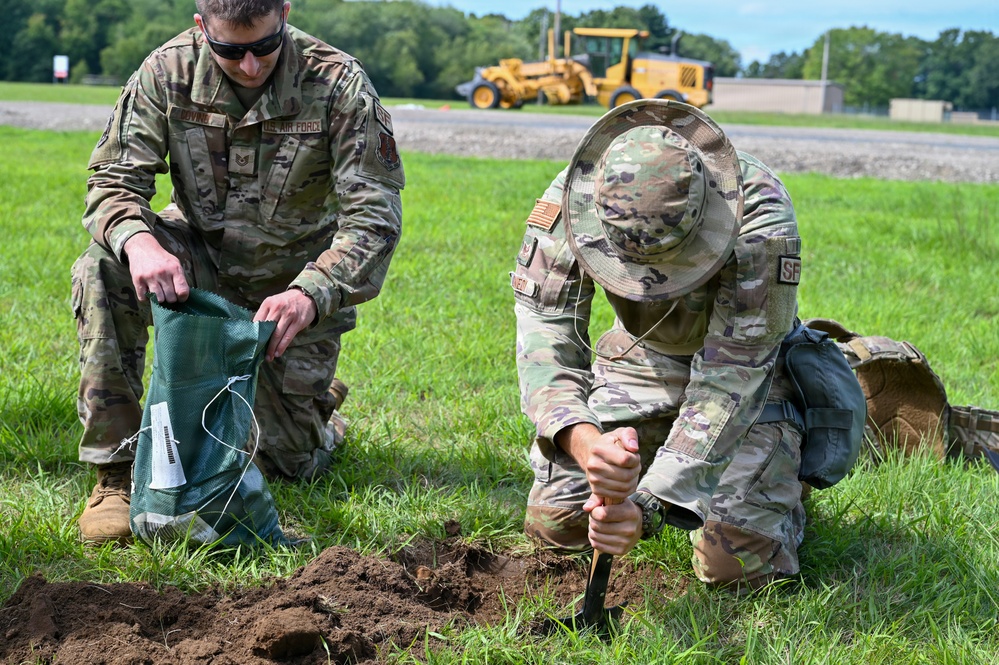 Two U.S. Air Force personnel in OCP (Operational Camouflage Pattern) uniforms are kneeling on grass, filling a sandbag with soil. One airman holds open a green sandbag while the other uses a hand tool to dig and transfer dirt. A yellow construction vehicle is visible in the background on a paved surface.