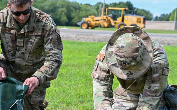 Two U.S. Air Force personnel in OCP (Operational Camouflage Pattern) uniforms are kneeling on grass, filling a sandbag with soil. One airman holds open a green sandbag while the other uses a hand tool to dig and transfer dirt. A yellow construction vehicle is visible in the background on a paved surface.