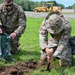 Two U.S. Air Force personnel in OCP (Operational Camouflage Pattern) uniforms are kneeling on grass, filling a sandbag with soil. One airman holds open a green sandbag while the other uses a hand tool to dig and transfer dirt. A yellow construction vehicle is visible in the background on a paved surface.