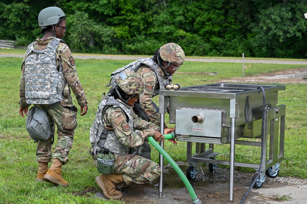 Three military personnel in camouflage uniforms and body armor are working with a stainless steel field equipment unit outdoors on a grassy area. Two soldiers are actively engaged with the equipment, with one kneeling and connecting a green flexible hose to the unit labeled 'HOT EXHAUST', while a third soldier observes. The setting appears to be a military training exercise or field operation.