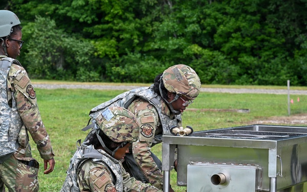 Three military personnel in camouflage uniforms and body armor are working with a stainless steel field equipment unit outdoors on a grassy area. Two soldiers are actively engaged with the equipment, with one kneeling and connecting a green flexible hose to the unit labeled 'HOT EXHAUST', while a third soldier observes. The setting appears to be a military training exercise or field operation.