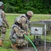 Three military personnel in camouflage uniforms and body armor are working with a stainless steel field equipment unit outdoors on a grassy area. Two soldiers are actively engaged with the equipment, with one kneeling and connecting a green flexible hose to the unit labeled 'HOT EXHAUST', while a third soldier observes. The setting appears to be a military training exercise or field operation.