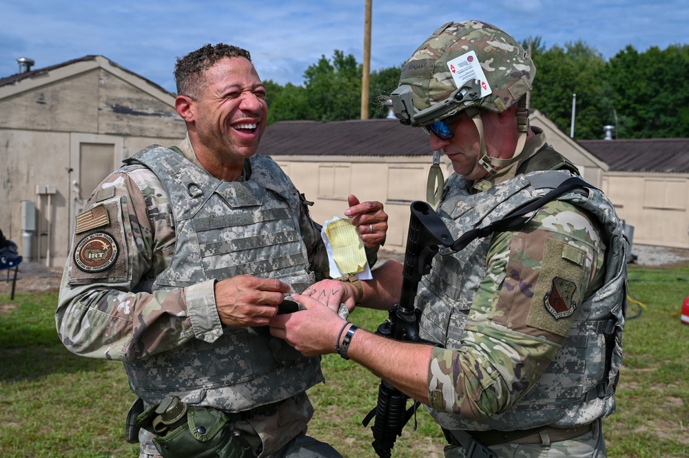 Two military personnel in camouflage uniforms are interacting outdoors at what appears to be a military installation or training facility. One soldier is laughing broadly while the other, wearing a helmet with mounted equipment, appears to be writing on or examining the first soldier's wrist. The interaction appears informal and lighthearted in nature.