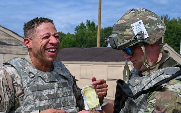 Two military personnel in camouflage uniforms are interacting outdoors at what appears to be a military installation or training facility. One soldier is laughing broadly while the other, wearing a helmet with mounted equipment, appears to be writing on or examining the first soldier's wrist. The interaction appears informal and lighthearted in nature.