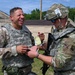 Two military personnel in camouflage uniforms are interacting outdoors at what appears to be a military installation or training facility. One soldier is laughing broadly while the other, wearing a helmet with mounted equipment, appears to be writing on or examining the first soldier's wrist. The interaction appears informal and lighthearted in nature.