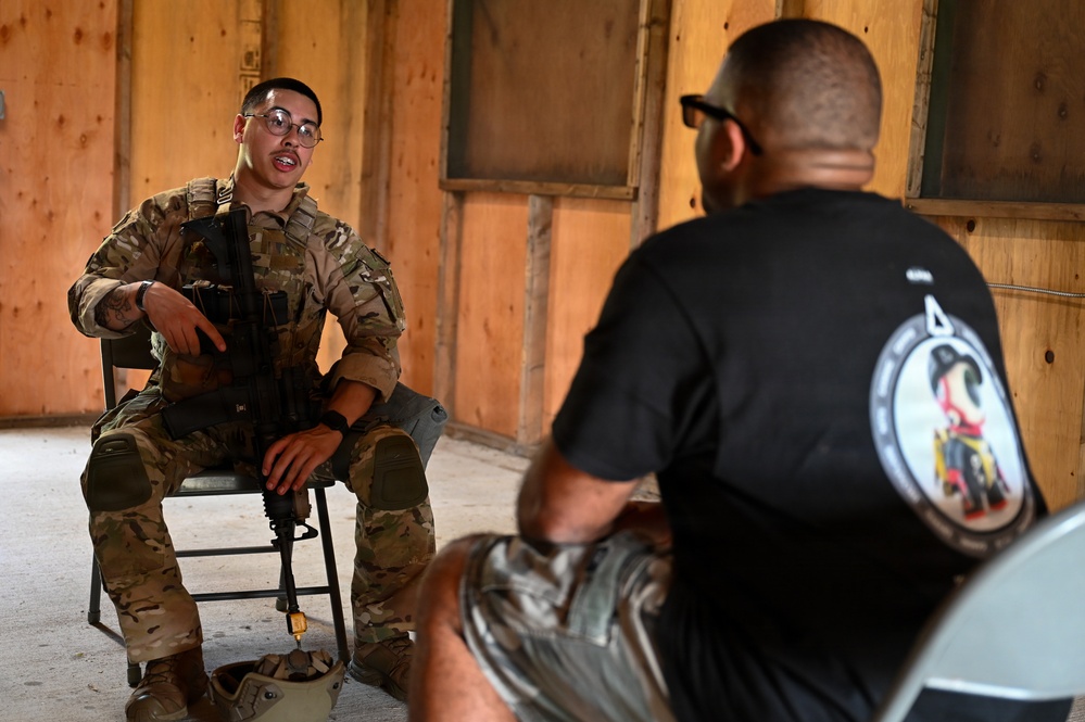 A soldier in full camouflage uniform and glasses sits on a folding chair holding a rifle while speaking with a civilian or off-duty personnel wearing a black t-shirt with a unit patch. The setting appears to be inside a wooden structure, possibly a training facility or temporary building. The interaction suggests a briefing, debrief, or interview scenario.