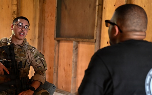 A soldier in full camouflage uniform and glasses sits on a folding chair holding a rifle while speaking with a civilian or off-duty personnel wearing a black t-shirt with a unit patch. The setting appears to be inside a wooden structure, possibly a training facility or temporary building. The interaction suggests a briefing, debrief, or interview scenario.