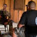 A soldier in full camouflage uniform and glasses sits on a folding chair holding a rifle while speaking with a civilian or off-duty personnel wearing a black t-shirt with a unit patch. The setting appears to be inside a wooden structure, possibly a training facility or temporary building. The interaction suggests a briefing, debrief, or interview scenario.