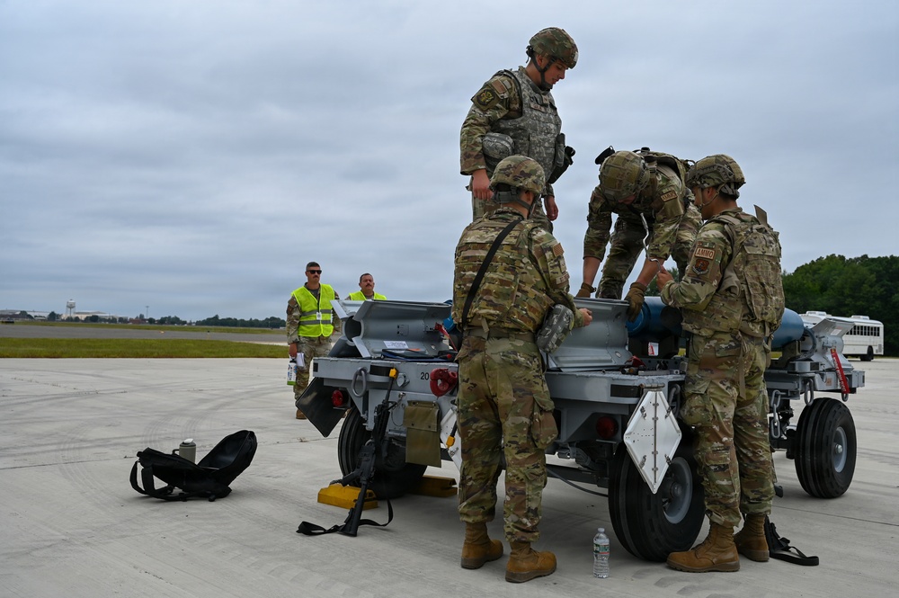 A group of military personnel in MultiCam/OCP camouflage uniforms are working around a munitions cart or weapons loading trailer on an airfield tarmac. Several soldiers wearing 'AMMO' patches are handling equipment on the cart, while two civilian or support personnel in high-visibility yellow vests observe in the background. The scene appears to depict an ammunition handling or weapons loading operation at a military airfield.
