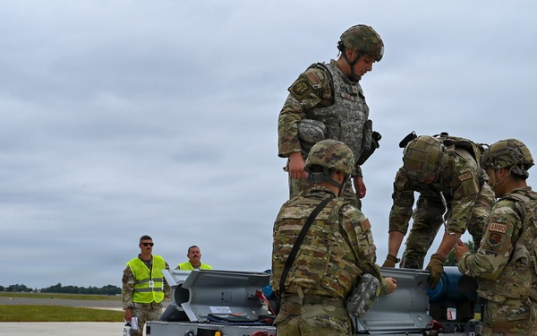 A group of military personnel in MultiCam/OCP camouflage uniforms are working around a munitions cart or weapons loading trailer on an airfield tarmac. Several soldiers wearing 'AMMO' patches are handling equipment on the cart, while two civilian or support personnel in high-visibility yellow vests observe in the background. The scene appears to depict an ammunition handling or weapons loading operation at a military airfield.