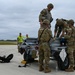 A group of military personnel in MultiCam/OCP camouflage uniforms are working around a munitions cart or weapons loading trailer on an airfield tarmac. Several soldiers wearing 'AMMO' patches are handling equipment on the cart, while two civilian or support personnel in high-visibility yellow vests observe in the background. The scene appears to depict an ammunition handling or weapons loading operation at a military airfield.