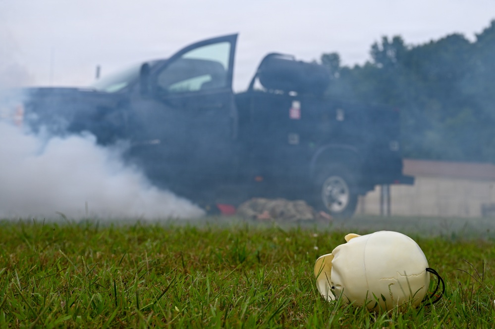A training or simulation scenario depicting a vehicle fire or explosion drill. A dark pickup truck with its door open is emitting heavy smoke in the background, while a white CPR/medical training mannequin head lies on the grass in the foreground. The scene appears to be a first responder or emergency services training exercise.