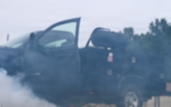 A training or simulation scenario depicting a vehicle fire or explosion drill. A dark pickup truck with its door open is emitting heavy smoke in the background, while a white CPR/medical training mannequin head lies on the grass in the foreground. The scene appears to be a first responder or emergency services training exercise.