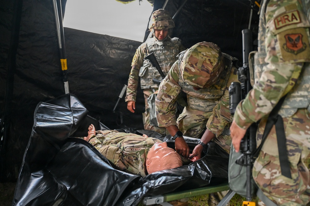 Military personnel in OCP (Operational Camouflage Pattern) uniforms are conducting a medical training exercise involving a mannequin casualty on a field litter/stretcher inside a tactical tent. One soldier is leaning over the mannequin performing a simulated medical assessment or treatment. A patch reading 'HRA' and an Air Force wing insignia are visible on one soldier's uniform, suggesting joint-service participation.