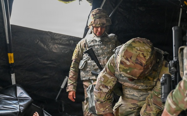 Military personnel in OCP (Operational Camouflage Pattern) uniforms are conducting a medical training exercise involving a mannequin casualty on a field litter/stretcher inside a tactical tent. One soldier is leaning over the mannequin performing a simulated medical assessment or treatment. A patch reading 'HRA' and an Air Force wing insignia are visible on one soldier's uniform, suggesting joint-service participation.