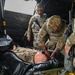 Military personnel in OCP (Operational Camouflage Pattern) uniforms are conducting a medical training exercise involving a mannequin casualty on a field litter/stretcher inside a tactical tent. One soldier is leaning over the mannequin performing a simulated medical assessment or treatment. A patch reading 'HRA' and an Air Force wing insignia are visible on one soldier's uniform, suggesting joint-service participation.