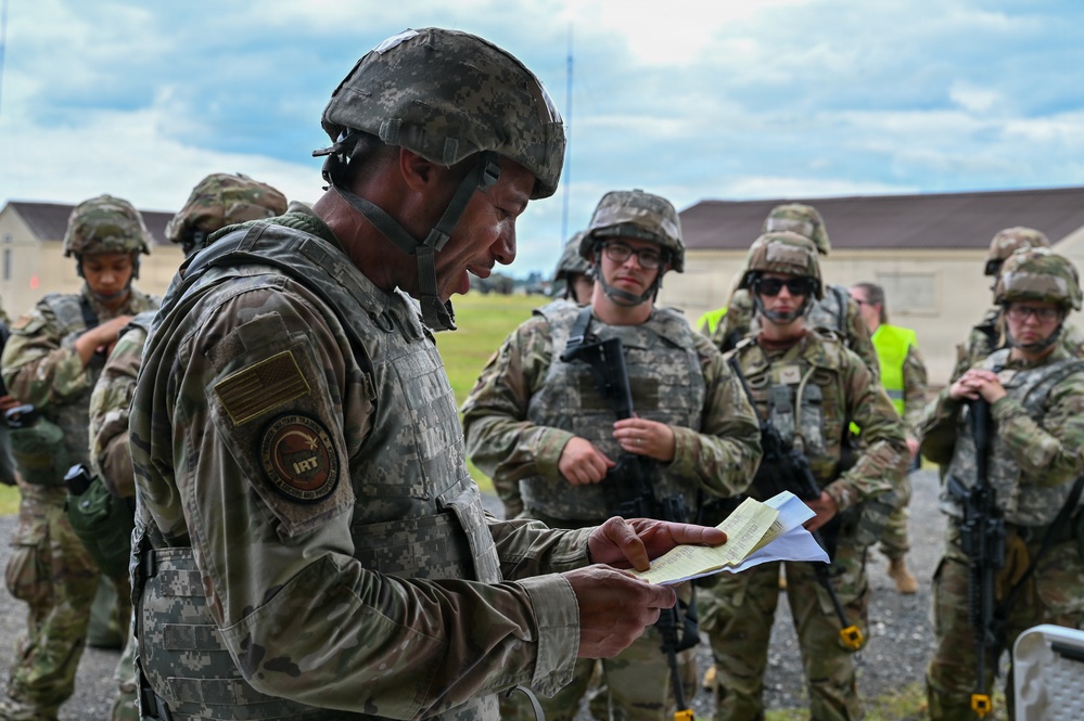 A U.S. Army soldier in the foreground, wearing a combat helmet and IRT (Instructor/Role Player Team) patch, reviews documents while a group of soldiers in OCP (Operational Camouflage Pattern) uniforms stand attentively in the background. The setting appears to be a military training installation, with barracks-style buildings visible in the background. This appears to be a training exercise briefing or after-action review.