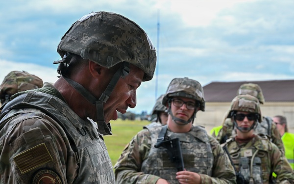 A U.S. Army soldier in the foreground, wearing a combat helmet and IRT (Instructor/Role Player Team) patch, reviews documents while a group of soldiers in OCP (Operational Camouflage Pattern) uniforms stand attentively in the background. The setting appears to be a military training installation, with barracks-style buildings visible in the background. This appears to be a training exercise briefing or after-action review.
