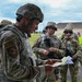 A U.S. Army soldier in the foreground, wearing a combat helmet and IRT (Instructor/Role Player Team) patch, reviews documents while a group of soldiers in OCP (Operational Camouflage Pattern) uniforms stand attentively in the background. The setting appears to be a military training installation, with barracks-style buildings visible in the background. This appears to be a training exercise briefing or after-action review.