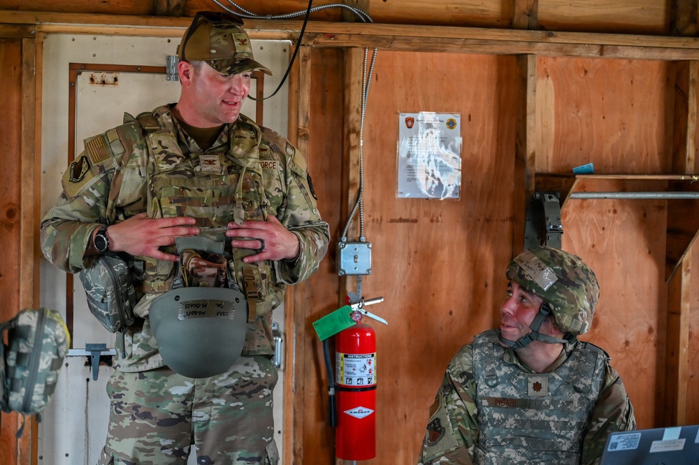 Two military personnel in camouflage uniforms are inside a wooden structure, appearing to be in conversation. The standing soldier is wearing OCP (Operational Camouflage Pattern) uniform and holding a combat helmet, while the seated soldier wears ACU (Army Combat Uniform) pattern with a helmet on. The setting appears to be a forward operating base or training facility.