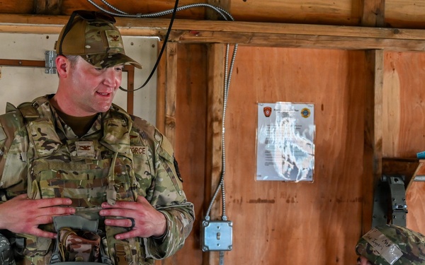Two military personnel in camouflage uniforms are inside a wooden structure, appearing to be in conversation. The standing soldier is wearing OCP (Operational Camouflage Pattern) uniform and holding a combat helmet, while the seated soldier wears ACU (Army Combat Uniform) pattern with a helmet on. The setting appears to be a forward operating base or training facility.