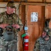 Two military personnel in camouflage uniforms are inside a wooden structure, appearing to be in conversation. The standing soldier is wearing OCP (Operational Camouflage Pattern) uniform and holding a combat helmet, while the seated soldier wears ACU (Army Combat Uniform) pattern with a helmet on. The setting appears to be a forward operating base or training facility.