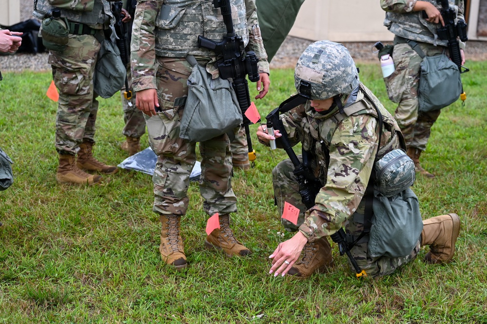 Military personnel in camouflage uniforms are conducting what appears to be a field training exercise on a grassy area. One soldier is kneeling on the ground examining or placing small orange marker flags, while holding a device in their other hand. Several other soldiers stand nearby observing or participating in the exercise.