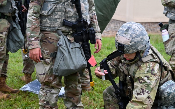 Military personnel in camouflage uniforms are conducting what appears to be a field training exercise on a grassy area. One soldier is kneeling on the ground examining or placing small orange marker flags, while holding a device in their other hand. Several other soldiers stand nearby observing or participating in the exercise.