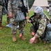 Military personnel in camouflage uniforms are conducting what appears to be a field training exercise on a grassy area. One soldier is kneeling on the ground examining or placing small orange marker flags, while holding a device in their other hand. Several other soldiers stand nearby observing or participating in the exercise.