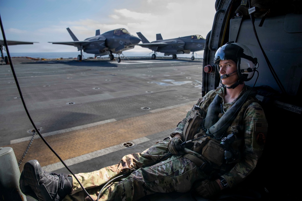 A military service member in full combat gear and flight helmet sits in the open doorway of a helicopter on the flight deck of a naval vessel. In the background, two F-35B Lightning II stealth fighter jets are parked on the deck. The scene suggests a joint military operation involving rotary and fixed-wing aircraft aboard an amphibious assault ship.