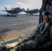 A military service member in full combat gear and flight helmet sits in the open doorway of a helicopter on the flight deck of a naval vessel. In the background, two F-35B Lightning II stealth fighter jets are parked on the deck. The scene suggests a joint military operation involving rotary and fixed-wing aircraft aboard an amphibious assault ship.