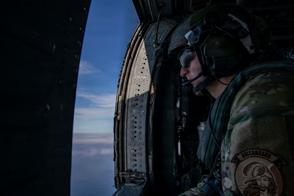 A military aviator or crew member is seated inside a helicopter, looking out through the open door at an aerial view of sky and clouds. The individual is wearing full flight gear including a helmet with visor, communications headset, and multicam camouflage uniform. A unit patch reading 'BUCCANEERS' with a pirate-style insignia is visible on the right shoulder sleeve.