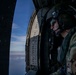 A military aviator or crew member is seated inside a helicopter, looking out through the open door at an aerial view of sky and clouds. The individual is wearing full flight gear including a helmet with visor, communications headset, and multicam camouflage uniform. A unit patch reading 'BUCCANEERS' with a pirate-style insignia is visible on the right shoulder sleeve.