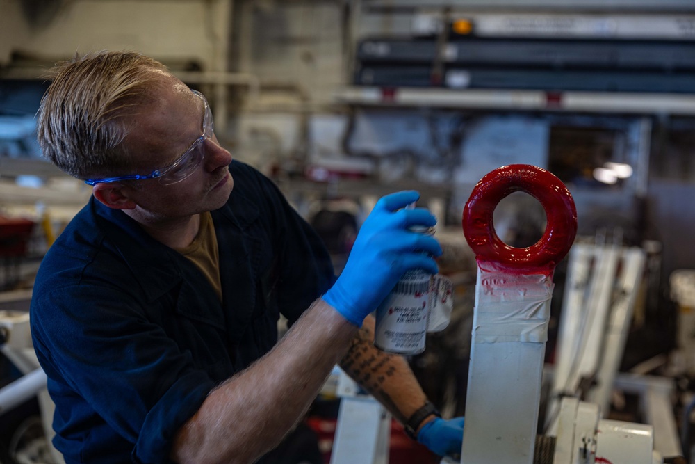 A male worker wearing blue nitrile gloves and safety glasses is spray painting a red circular ring/tow fitting on what appears to be white military or industrial equipment. The setting appears to be an industrial workspace or military vessel hangar bay. The worker is using a spray can to apply red paint to the circular component.