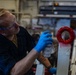 A male worker wearing blue nitrile gloves and safety glasses is spray painting a red circular ring/tow fitting on what appears to be white military or industrial equipment. The setting appears to be an industrial workspace or military vessel hangar bay. The worker is using a spray can to apply red paint to the circular component.