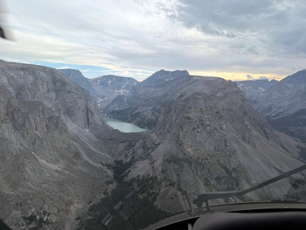 An aerial photograph taken from inside a small aircraft or helicopter cockpit, showing a dramatic mountain valley with a turquoise glacial lake nestled between steep rocky cliffs. The scene captures rugged, high-altitude terrain with patches of snow, sparse coniferous trees in the lower valley, and a partly cloudy sky with warm sunset tones on the horizon. The cockpit frame and instrument panel are partially visible in the foreground.