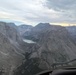 An aerial photograph taken from inside a small aircraft or helicopter cockpit, showing a dramatic mountain valley with a turquoise glacial lake nestled between steep rocky cliffs. The scene captures rugged, high-altitude terrain with patches of snow, sparse coniferous trees in the lower valley, and a partly cloudy sky with warm sunset tones on the horizon. The cockpit frame and instrument panel are partially visible in the foreground.