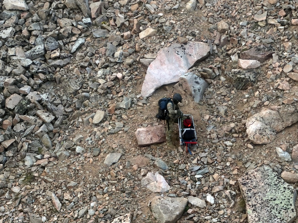 An aerial or elevated view of a rocky, boulder-strewn terrain showing a single individual with a large backpack standing near what appears to be a wheeled cart or litter. The person appears to be on a rugged mountain or scree field, surrounded by rocks of varying sizes. The scene suggests a backcountry expedition, rescue operation, or equipment transport in a remote rocky environment.