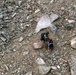 An aerial or elevated view of a rocky, boulder-strewn terrain showing a single individual with a large backpack standing near what appears to be a wheeled cart or litter. The person appears to be on a rugged mountain or scree field, surrounded by rocks of varying sizes. The scene suggests a backcountry expedition, rescue operation, or equipment transport in a remote rocky environment.