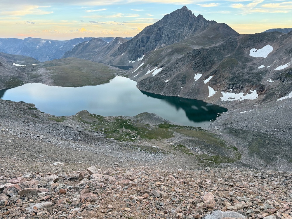 A high-altitude alpine landscape photographed from an elevated rocky vantage point, showing a calm reflective lake nestled in a glacially carved valley. A jagged rocky peak dominates the background center, with patches of snow visible on surrounding slopes. The image appears to be taken at either dawn or dusk based on the warm pastel sky tones.