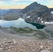 A high-altitude alpine landscape photographed from an elevated rocky vantage point, showing a calm reflective lake nestled in a glacially carved valley. A jagged rocky peak dominates the background center, with patches of snow visible on surrounding slopes. The image appears to be taken at either dawn or dusk based on the warm pastel sky tones.