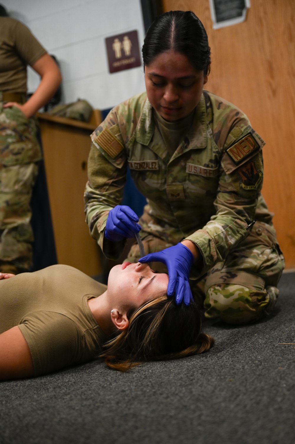 A U.S. Air Force medical personnel member wearing OCP (Operational Camouflage Pattern) uniform and blue nitrile gloves is performing a medical training exercise on a simulated casualty lying on the floor. The provider, whose name tape reads 'GONZALEZ,' appears to be conducting an airway assessment or nasopharyngeal airway (NPA) insertion procedure on a person lying supine. A second uniformed individual is visible in the background, suggesting a group training scenario.