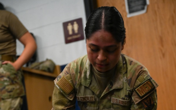A U.S. Air Force medical personnel member wearing OCP (Operational Camouflage Pattern) uniform and blue nitrile gloves is performing a medical training exercise on a simulated casualty lying on the floor. The provider, whose name tape reads 'GONZALEZ,' appears to be conducting an airway assessment or nasopharyngeal airway (NPA) insertion procedure on a person lying supine. A second uniformed individual is visible in the background, suggesting a group training scenario.